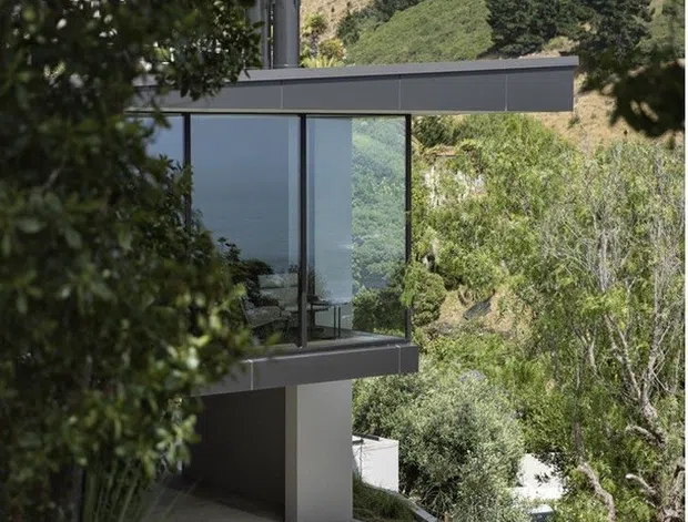 A contemporary multi-level house set into a densely vegetated, sloping landscape. The visible facade features a crisp flat roof, dark horizontal fascia, expansive floor-to-ceiling glazing with slim dark frames, and a projecting glass-fronted upper level supported by a solid column. Concrete terraces and retaining elements step down through the site, with mature trees and hillside vegetation surrounding the building.