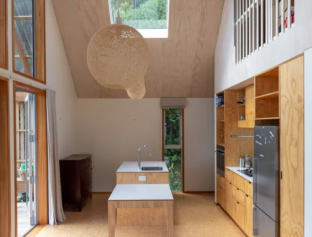 A bright, double-height kitchen viewed from an open living area, featuring extensive warm plywood cabinetry, a central island with an integrated sink, cork-toned flooring, tall glazing, and a large roof skylight. A timber slatted balustrade overlooks the kitchen from an upper level, while a woven pendant light and patterned rug add warmth and texture.