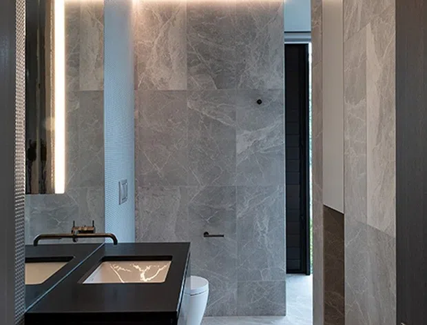 A contemporary bathroom viewed through an open doorway, featuring large-format grey marble-look wall and floor tiles, a floating dark vanity with integrated basins, a wall-hung toilet, black fixtures, and concealed linear LED lighting along the ceiling and wall junctions. The space has a narrow, elongated layout with a sleek, high-contrast finish.