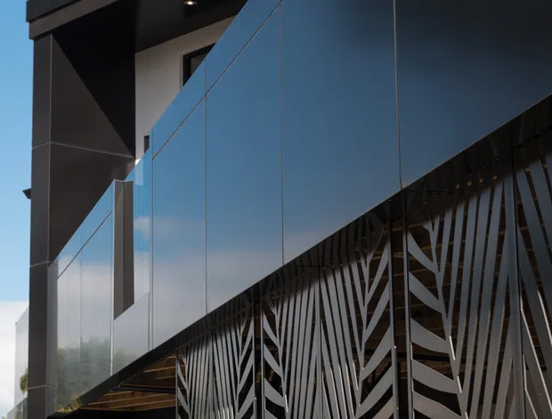 A close-up exterior view of a contemporary building featuring dark reflective cladding, sharp cantilevered roof forms, and a decorative metal privacy screen with an organic leaf-like pattern. Strong geometric lines, black and charcoal finishes, and reflective glazing dominate the facade against a clear blue sky.