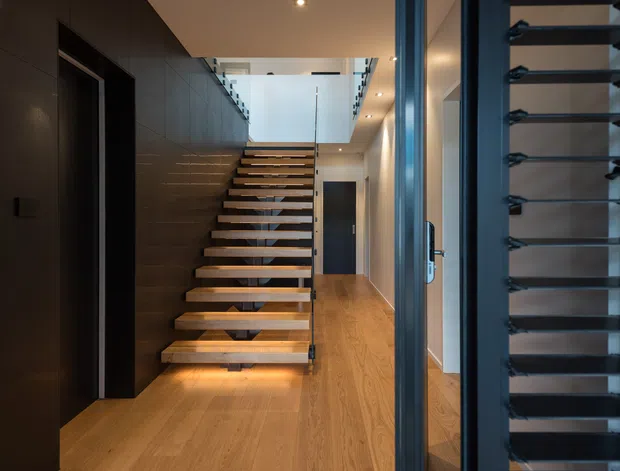 Interior view of a contemporary stair hall with floating timber treads, dark metal supports and handrails, and warm timber flooring. Dark built-in wall panelling and cabinetry line the left side, while a glazed and louvred dark-framed opening is visible on the right. Recessed lighting and a double-height stair void create a refined, modern atmosphere.