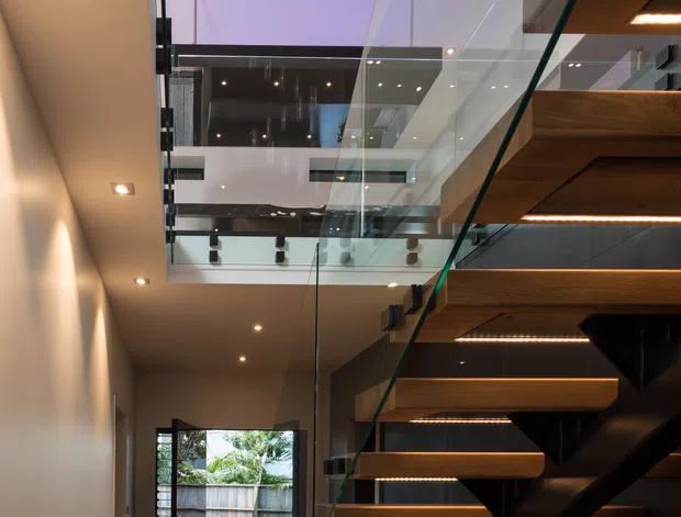 A tall contemporary interior hallway dominated by a floating timber staircase with dark structural supports, clear glass balustrades, and integrated LED lighting beneath each tread. The stairway rises through a multi-level void with glass-edged landings, recessed ceiling lights, and suspended cylindrical pendant lights. Warm timber flooring and stair treads contrast with white walls and dark joinery, while an open doorway leads outside at the end of the hall.