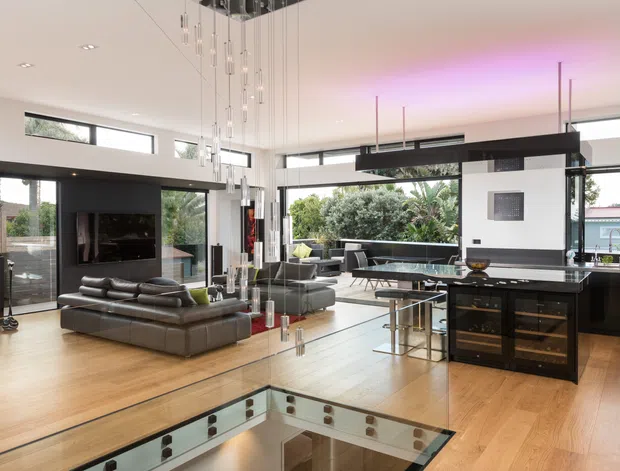 Open-plan contemporary living, dining and kitchen space with expansive glazing, warm timber flooring, a dark kitchen island and bar cabinetry, a sunken stair opening in the foreground, and a dramatic cluster of suspended pendant lights. The room has a bright, airy feel with subtle purple cove lighting and views to greenery outside.
