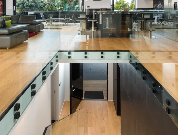 A contemporary open-plan living area viewed from an upper-level landing, with a dramatic stairwell descending through the centre. The space features warm timber flooring and stairs, extensive black-framed glazing, glass balustrades, dark built-in cabinetry, grey sectional seating, and a cluster of slender pendant lights. Lush garden planting is visible through the windows, while a kitchen or bar area sits beyond the living zone.