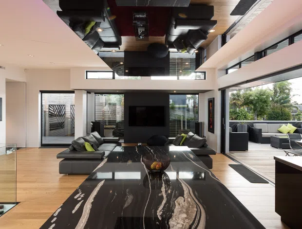 A dramatic contemporary open-plan living area viewed from a kitchen island. The foreground features a large black marble island benchtop with striking white veining and dark timber cabinetry. Beyond it is a sophisticated lounge with black sofas, a central television wall, extensive glazing, timber flooring, and a mirrored ceiling detail reflecting the room and pendant lighting. The space opens toward an outdoor seating area and garden.