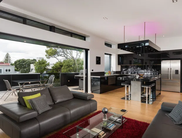 Open-plan contemporary living, dining and kitchen interior with polished dark cabinetry, a large black island, stainless-steel appliances, timber flooring and expansive sliding glass doors opening to a terrace. Dark leather sofas and a red rug define the living area, while a black-framed clerestory and recessed ceiling lighting add architectural detail. The space has a strong indoor-outdoor connection and overlooks neighbouring greenery and rooftops.