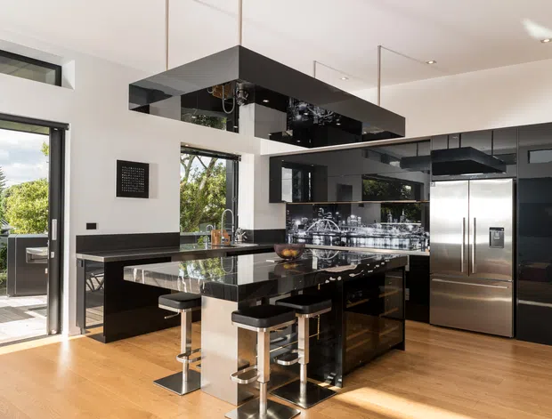 A contemporary open-plan kitchen with a glossy black island, integrated black cabinetry, reflective overhead rangehood, stainless-steel appliances, and bar seating. Warm timber flooring contrasts with white walls and large glazed openings that bring in abundant daylight and views of greenery.