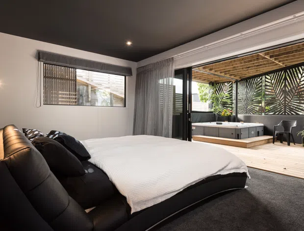A contemporary bedroom with a large black upholstered bed, white bedding, dark carpet, charcoal ceiling, and sheer floor-to-ceiling curtains. Sliding glass doors open directly onto a timber-decked outdoor terrace featuring a spa, outdoor seating, timber overhead screening, and patterned privacy panels.