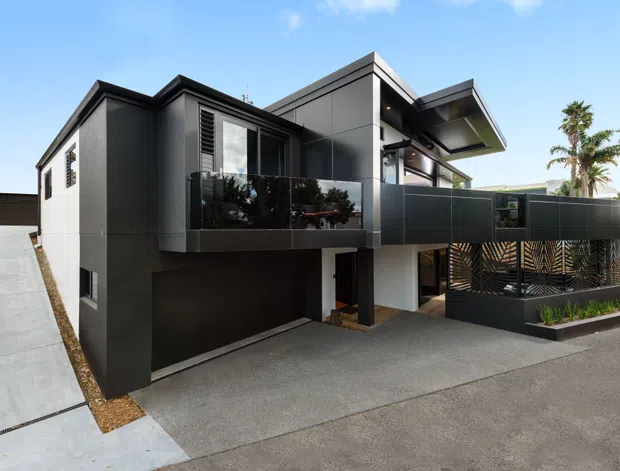 Exterior view of a contemporary multi-level residence with dark charcoal cladding, projecting box-like forms, angled roof planes, upper-level balconies with dark privacy screens, and an integrated ground-level garage. The home fronts a paved driveway and includes tropical palms visible in the background.
