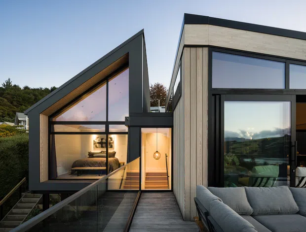 Contemporary multi-volume house exterior at dusk, featuring dramatic steep gabled glazing, dark metal cladding, pale vertical timber cladding, and expansive black-framed windows. A narrow elevated timber walkway connects the glazed bedroom wing to the main volume, while a partially visible outdoor lounge with a grey sofa occupies the foreground. Warm interior lighting contrasts with the cool blue exterior light, and surrounding vegetation and distant landscape are visible beyond the house.