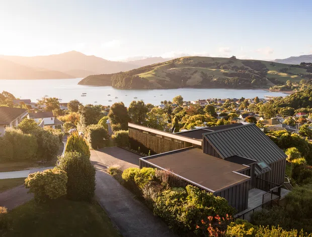 Aerial view of a modern dark-clad residential building on a landscaped hillside, overlooking a sheltered bay, coastal settlement, and steep green hills. The home has a low-slung flat-roofed section paired with a steep pitched metal roof, extensive glazing, and carefully planted gardens.