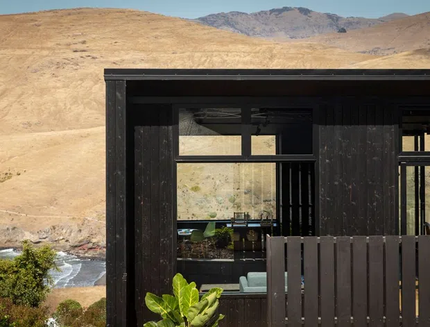Exterior view of a dark, timber-clad modern building set against a dry mountainous landscape, with large glazed openings revealing part of the interior and a dark vertical-slat privacy screen or balcony enclosure in the foreground. A river or stream is visible below in the surrounding terrain.