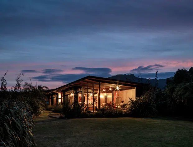 A single-storey modern pavilion-style house photographed at dusk, set within a landscaped garden. The low-pitched roof, extensive warm-lit glazing, timber-framed structure and open terrace create a strong indoor-outdoor connection, with mountains visible in the background.