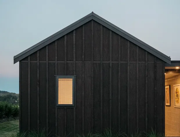 A small, dark-clad gable-roofed building viewed externally at dusk. The facade features vertical timber-like boards, a small shuttered window, and a partially visible warmly lit entrance on the right. Native-style grasses and low planting surround the structure, with a hilly landscape faintly visible in the background.