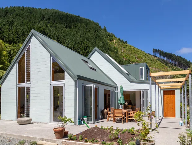 Exterior view of a light-coloured, multi-gabled home set against a wooded hillside. The house has steep pitched metal roofs, white rendered or painted masonry walls, extensive glazing, a sheltered outdoor dining patio, and a timber pergola over the entry walkway. A small landscaped garden and concrete paving surround the building.