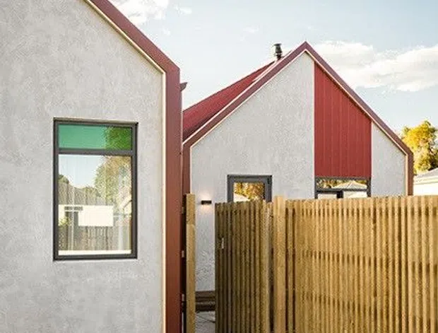 Exterior view of a contemporary residential building composed of compact gabled volumes. The facades are light grey textured render with contrasting red vertical metal cladding and red roof-edge trims. A timber privacy fence and gate line a narrow concrete-paved walkway, with a small lawn in the foreground. The image is photographed in bright daylight and emphasizes the building envelope and pedestrian entry.