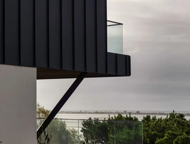 Exterior architectural view of a contemporary coastal home, featuring dark vertical cladding, a projecting upper volume supported by a diagonal steel brace, a glass-balustraded timber deck, and landscaped vegetation. The ocean and shoreline are visible in the distance beneath an overcast sky.