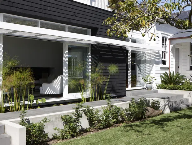 Exterior view of a contemporary residential extension with dark horizontal cladding, crisp white framing, large glazed sliding doors, and a landscaped stepped front garden. The open living area is visible through the glazing, while the original light-coloured house remains evident to the right. Strong sunlight creates patterned shadows across the facade and terrace.