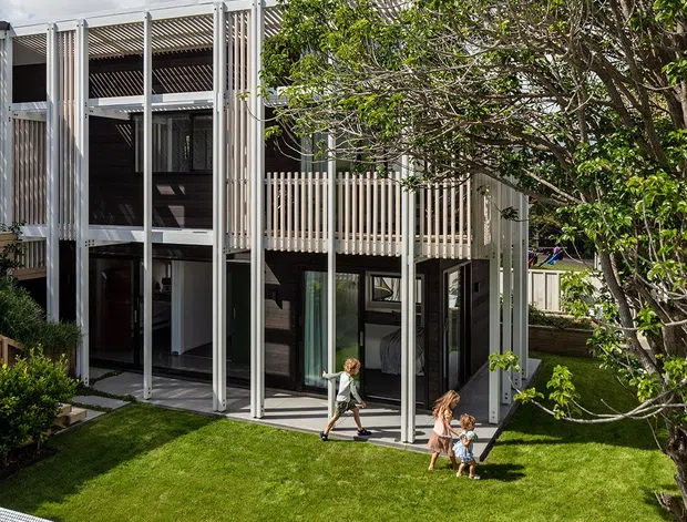 Exterior view of a contemporary multi-level residential home with extensive glazing, white vertical structural fins, screened upper-level terraces, and dark interior surfaces. The house opens onto a manicured lawn beside a tiled swimming pool, with mature landscaping and a large tree in the foreground. Bright daylight and partly cloudy sky illuminate the modern facade.
