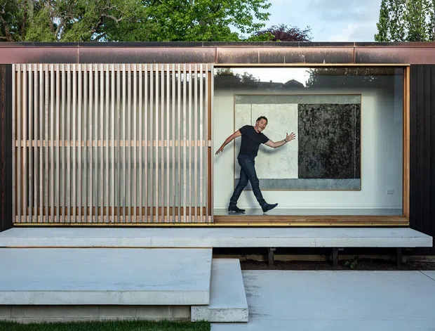 A contemporary single-storey building facade with dark vertical cladding, a broad rust-toned metal roof edge, and a large glazed opening. Vertical timber privacy screens flank the glazing, while a pale concrete platform and stepped entry extend across the foreground. Through the window, a person and large monochromatic artwork are visible inside. Mature trees appear behind the building.
