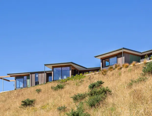 Exterior view of a contemporary multi-volume house stepping along a dry, sloping site. The building features flat, shallow-pitched roof planes, extensive floor-to-ceiling glazing, dark window frames, timber-toned vertical cladding, and projecting covered outdoor areas. Native-looking grasses and shrubs surround the structure beneath a clear blue sky.