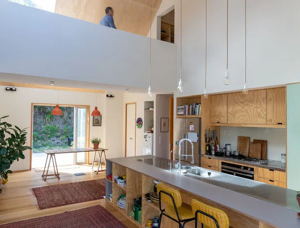 Bright open-plan kitchen and living space with a large island and raised breakfast bar, timber cabinetry, stainless-steel benchtops, and three bar stools. The room has a dramatic double-height ceiling lined with warm plywood, exposed pendant lights, a mezzanine or upper-level opening, timber floors, patterned rugs, and large glazing opening toward greenery.