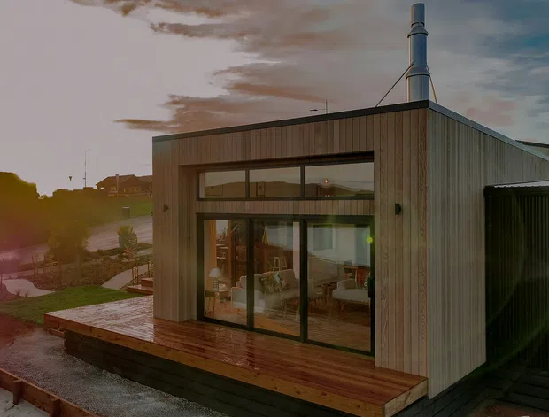 Exterior view of a compact contemporary timber-clad house or extension with a low-pitched flat roof, large black-framed glazed doors and clerestory windows, and a raised timber deck. Warm interior furnishings are visible through the glazing, while a tall metal flue rises from the roof. The image is photographed at dusk with wet decking and subdued natural light.