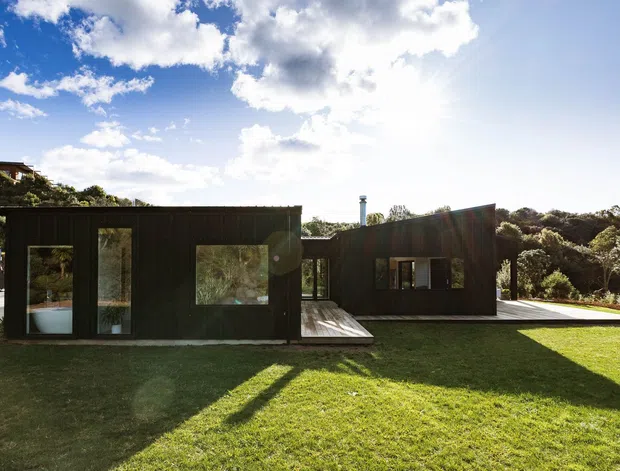 A low, dark-clad modern house set within a landscaped suburban or bushy site. The building is composed of simple rectilinear volumes with extensive glazing, a projecting timber deck, and a gently sloping roofline. A broad lawn occupies the foreground, while mature vegetation and a neighbouring hillside house sit behind it. Bright sunlight and a dramatic blue sky create strong shadows across the grass.
