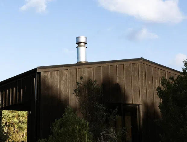 Exterior view of a compact, single-storey building clad in dark vertical timber boards, with a low pitched roof and a prominent metal chimney. Dense planting partially screens the facade, while the clear sky and simple geometric form create a restrained, contemporary appearance.