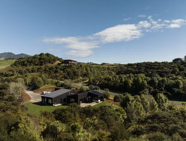 Aerial view of a low, dark-clad contemporary house set within a heavily vegetated, hilly rural landscape. The residence has a long low-pitched roof, several connected wings, and broad glazing, with a curved driveway and dense native-looking planting surrounding the site. Mountains and open countryside are visible in the distance beneath a bright blue sky.