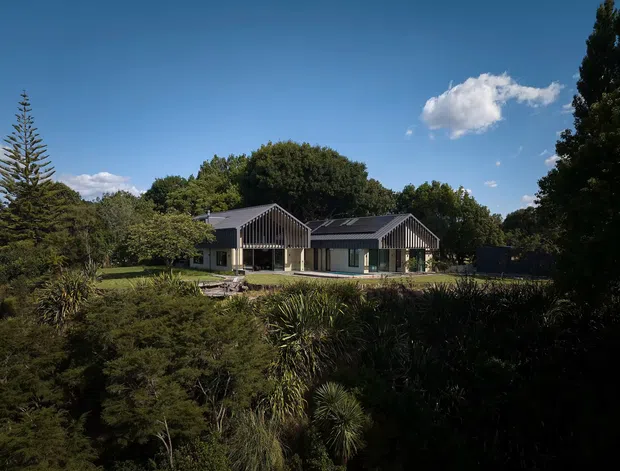 Exterior view of a low, contemporary single-storey house set within mature landscaped vegetation. The building has multiple pitched gable forms, dark roofing, light walls, and prominent vertical timber or metal screening at the gable ends. A pool or long reflective water feature appears in front of the central living area, with dense gardens surrounding the property under a clear blue sky.