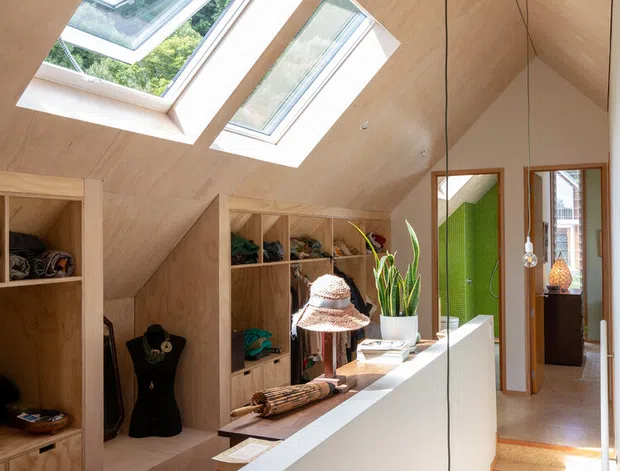 Bright contemporary attic dressing room or walk-in wardrobe with extensive custom plywood cabinetry, open shelving, a long white built-in counter, and a stair landing. The sloped timber-lined ceiling incorporates multiple skylights, while a full-height mirror reflects the space. Sunlight creates a warm, airy atmosphere, with indoor plants and clothing/display items adding lived-in detail.