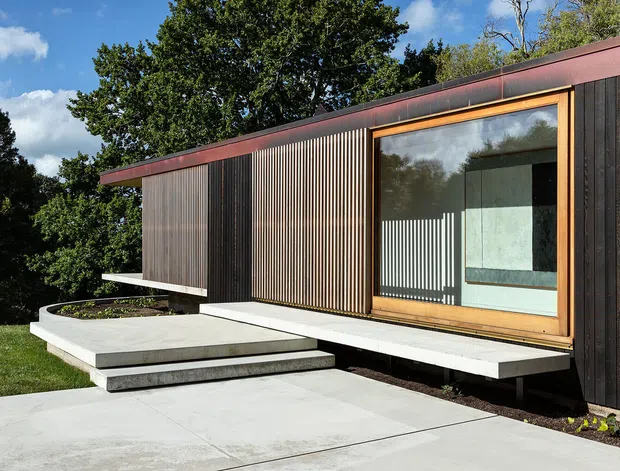 Exterior view of a compact, single-storey modern building set within a leafy landscape. The facade combines dark vertical timber cladding with finely spaced timber screening, a broad timber-framed glazed opening, and a low reddish-brown metal roof edge. A raised concrete terrace and stepped concrete entry path lead to the building, with the interior visible through the glazing.