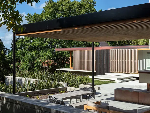 Contemporary residential exterior with a broad black flat-roofed covered terrace or carport-like canopy, timber slatted soffit, and slender dark steel posts. The paved outdoor space includes built-in concrete planters, low seating, and a dining setting, overlooking a modern single-storey pavilion clad in vertical dark timber slats and glazing. Lush established vegetation surrounds the structured hardscape.