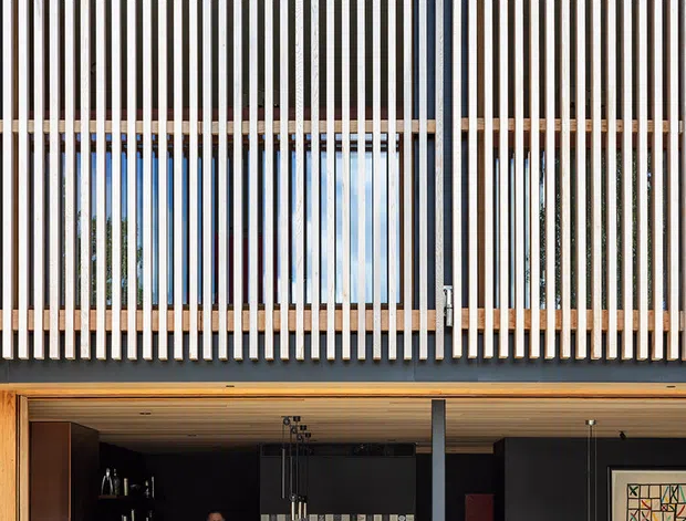 A contemporary two-storey facade with a dramatic vertical timber batten screen over large glazed openings. The lower level is an open-plan interior featuring a dark kitchen and bar area, central island seating, exposed dark structural columns, timber-lined ceiling, and an adjacent dining space. The upper facade combines warm timber framing with closely spaced vertical battens, allowing filtered views through the glazing.
