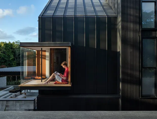 Exterior view of a contemporary dark-clad building with a steeply pitched, glazed roof volume and a projecting glass-enclosed reading nook. Vertical black cladding, tall narrow windows, and crisp geometric lines create a sculptural modern facade. Terraced concrete paving and low retaining walls sit in the foreground, with a person seated inside the glazed projection.
