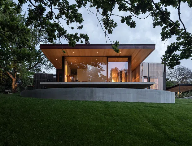 Exterior view of a low-slung contemporary house set within a landscaped, tree-lined site. The composition highlights a projecting flat roof with warm timber soffit, extensive floor-to-ceiling glazing, dark upper cladding, and a raised concrete terrace or retaining wall overlooking a sloping lawn.
