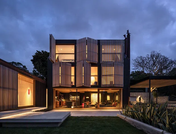 Twilight exterior view of a contemporary multi-storey residence arranged around a landscaped courtyard. The dark facade features extensive vertical timber or metal screening, large glazed openings, warm interior lighting, and projecting louvred panels. A lower side pavilion and covered outdoor structure frame the central house, with concrete paving and raised garden beds in the foreground.