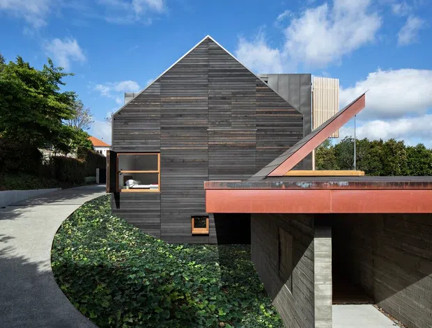 Exterior view of a contemporary gable-roofed building clad in horizontal weathered timber boards. A curved paved walkway leads beside landscaped groundcover to a recessed garage or covered entry, with a prominent rust-coloured metal roof or canopy element projecting across the foreground. The composition features strong geometric forms, a pitched gable facade, and contrasting vertical metal screening under a bright blue sky.