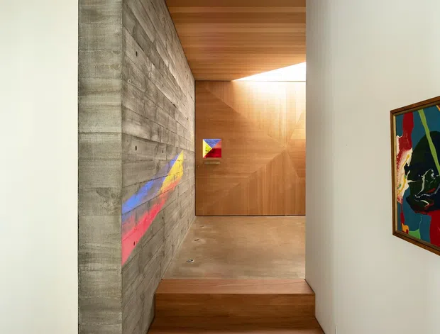 A refined contemporary interior stairway and landing with warm timber lining, a polished concrete wall and floor, and recessed lighting. The compact stair rises beside a textured concrete feature wall, leading to a timber-clad landing with a bright clerestory opening. Colourful abstract artworks provide small accents against the neutral architectural palette.