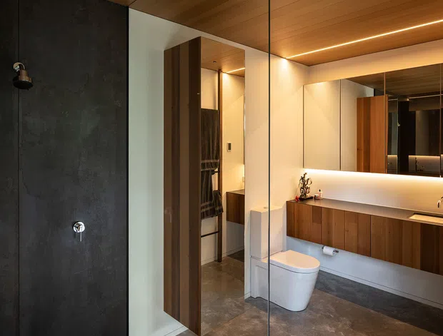 A refined contemporary bathroom featuring a dark textured shower wall enclosed by frameless glass, a wall-mounted timber vanity with a pale benchtop, a large mirrored splashback, warm timber ceiling lining, recessed linear lighting, and a white toilet. A timber door opens toward a partially visible adjoining space. The palette is warm, dark, and minimalist with integrated architectural lighting.