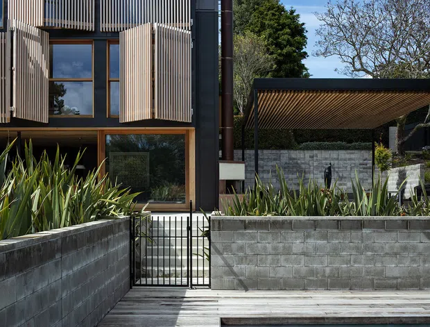 Contemporary multi-level residential exterior with dark cladding, extensive vertical timber screening, and warm timber-framed glazing. A timber pool deck and swimming pool occupy the foreground, while raised garden beds with architectural planting, concrete block retaining walls, a metal gate, and a covered outdoor structure frame the property. Bright daylight and strong linear shadows emphasize the facade and landscape geometry.