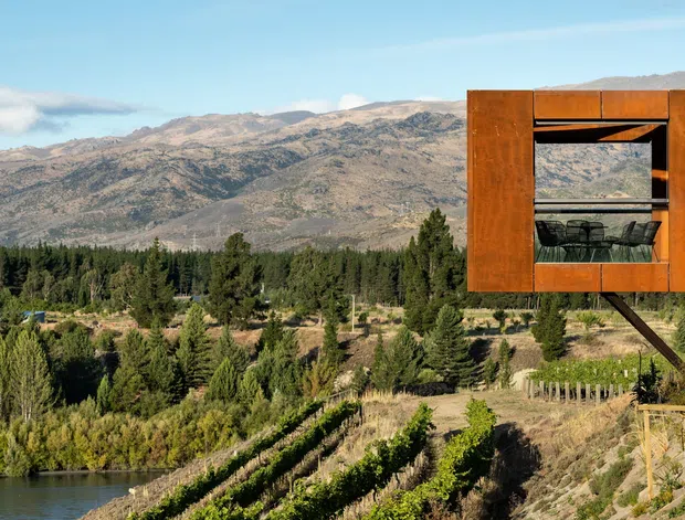 Exterior view of a compact elevated timber-clad structure projecting over a landscaped rural property, with a glazed opening revealing a small interior seating area. The building overlooks a lake or water edge, vineyards or planted rows, dense trees, and rugged mountains under a clear sky. The composition emphasises the dramatic natural setting and the warm rust-brown facade.