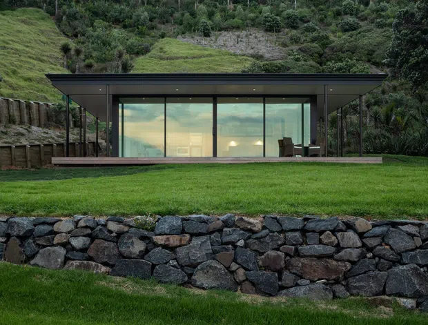 A low, single-storey modern pavilion set into a lush sloping landscape. The building has a thin flat roof, dark slender framing, extensive floor-to-ceiling glazing, and a narrow timber-toned deck. The interior is minimally visible through the glass, while the foreground features manicured lawn and a substantial dry-stacked dark stone retaining wall.