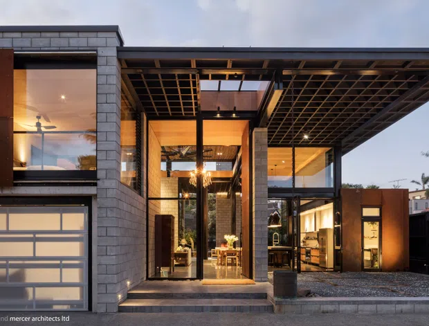 A contemporary multi-level residential facade viewed from the street at dusk. The home features grey masonry block walls, extensive floor-to-ceiling glazing, warm timber-lined interior ceilings, dark exposed steel framing, a broad projecting flat roof, and a translucent-panel garage door. A glazed entry courtyard and illuminated interiors are visible behind the facade, with gravel landscaping and a dark boundary fence.