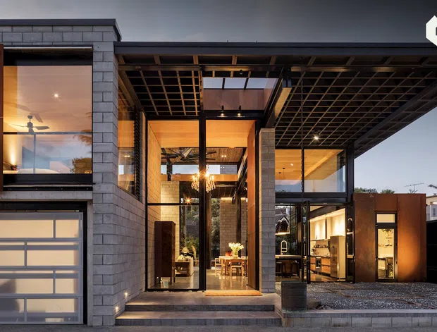 Exterior view of a contemporary multi-level residence at dusk, featuring a dark gridded roof canopy, exposed steel framing, extensive glazing, grey masonry block walls, and warm illuminated interiors. A translucent sectional garage door is visible at left, with a recessed entry and sheltered outdoor threshold at center.