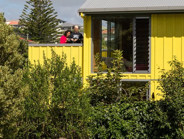 Exterior view of a compact, brightly painted yellow weatherboard house with a light grey corrugated metal gable roof. The facade features dark-framed glazing, a tall narrow louvred window, and simple decorative trim. Dense landscaped planting fills the foreground, with a yellow boundary wall and two people visible on an elevated outdoor area behind it. Other residential roofs and a tall conifer appear in the background.
