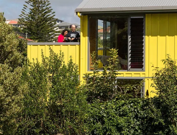 Exterior view of a bright yellow, single-storey residential house surrounded by dense garden planting. The facade has vertical weatherboard cladding, a pale corrugated pitched roof, large glazed windows, and dark external louvres. A small raised terrace or balcony with matching yellow balustrade is visible, with two people standing behind it. Neighbouring homes and a tall conifer appear in the background.