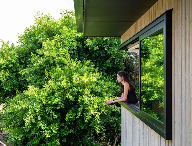 Exterior view of a contemporary timber-clad building with a projecting roof eave and a large open awning window. A person is seated at the window, looking out over dense green vegetation. The facade combines vertical light timber boards with dark framed glazing, creating a strong indoor–outdoor connection.