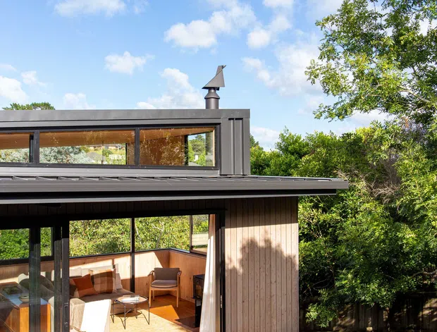 Exterior view of a compact contemporary timber-clad house opening onto a broad timber deck. Large black-framed glazed doors connect the deck to a warm living area, while a raised clerestory-style window band and a small roof vent add architectural detail. Dense vegetation surrounds the home, and the image is photographed in bright daylight beneath a partly cloudy sky.